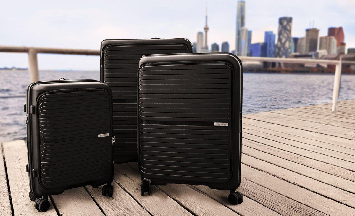 Three black Voyatique Drift hardside suitcase of different sizes arranged on a wooden dock with the blurred Toronto skyline and water in the background.